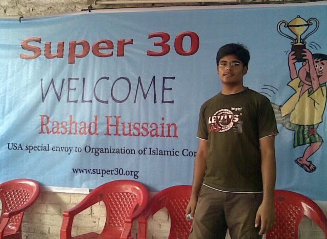 Archival photograph of a Super 30 event banner and a young man standing beside it.